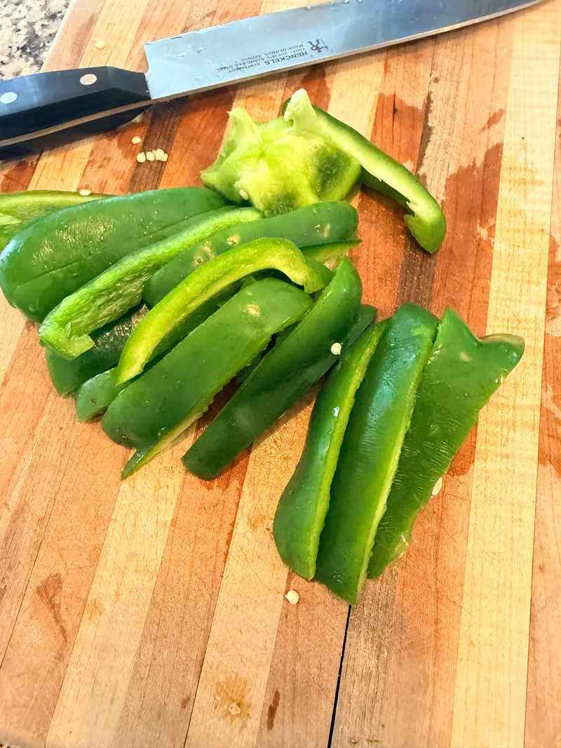 A green pepper sliced into strips on a wooden cutting board, knife alongside